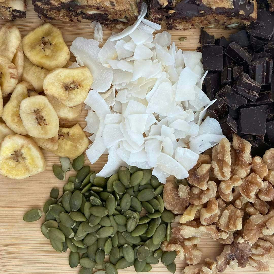Dried banana slices, coconut chips, chocolate chips, and nuts on a cutting board