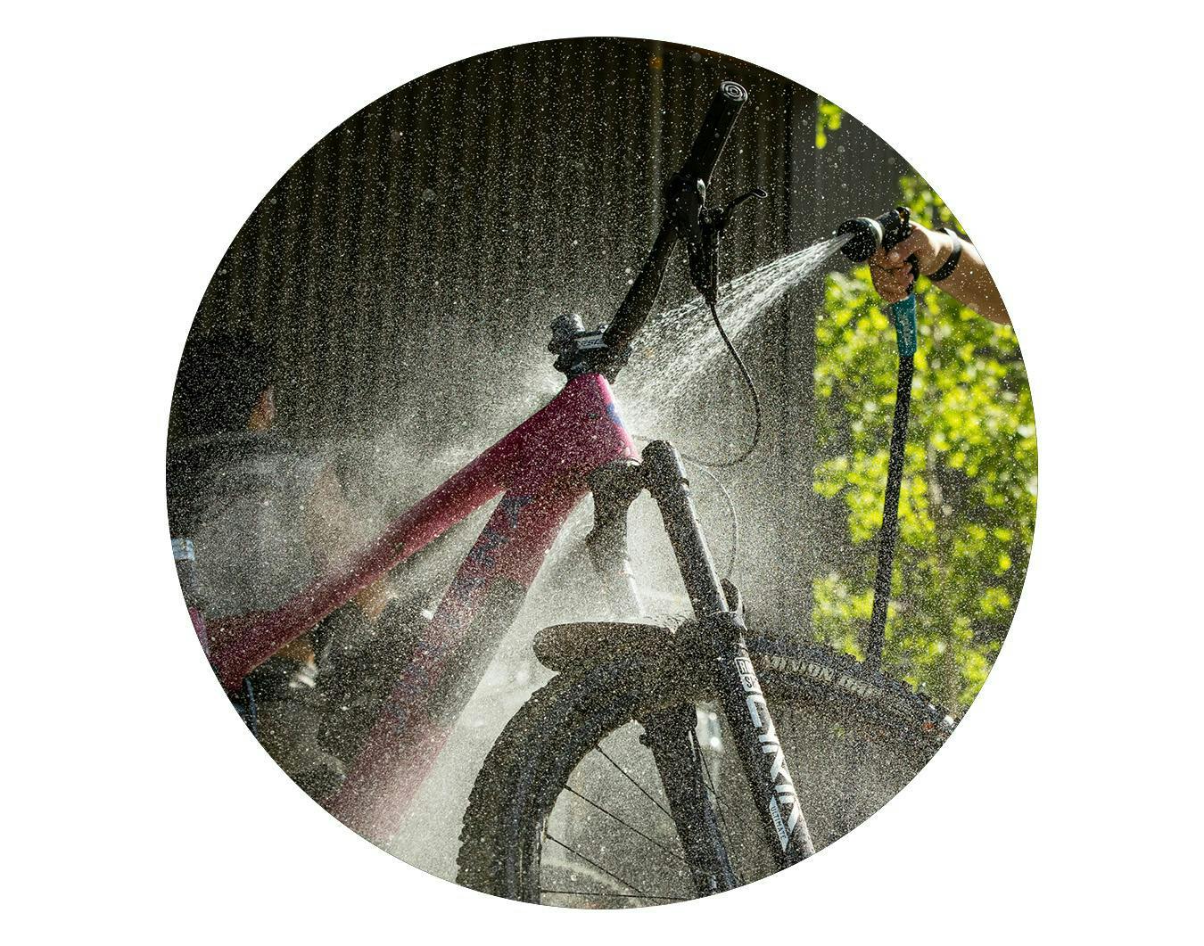 A circular image of someone cleaning their Juliana Bicycles Roubion with a hose and spray nozzle